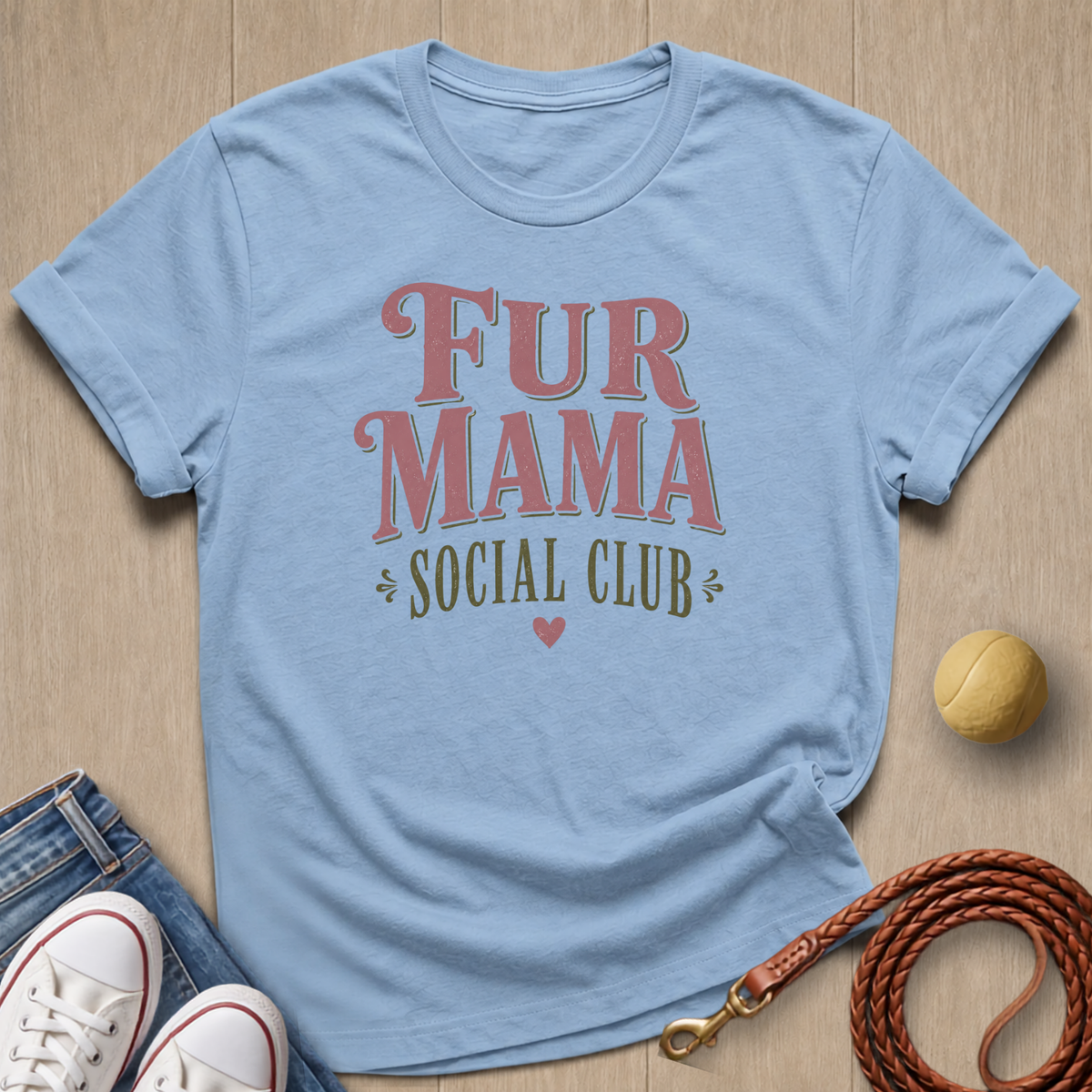 A light blue graphic t-shirt with 'FUR MAMA SOCIAL CLUB' printed on the front, styled in a flat-lay with jeans, white sneakers, a tennis ball, and a brown leather leash on a light wood background.